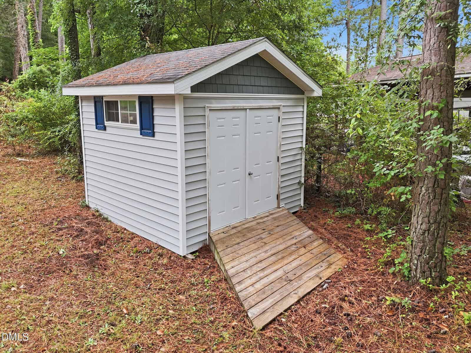 A small gray shed with double doors, blue shutters, and a ramp sits among pine trees and greenery. Pine needles cover the ground.