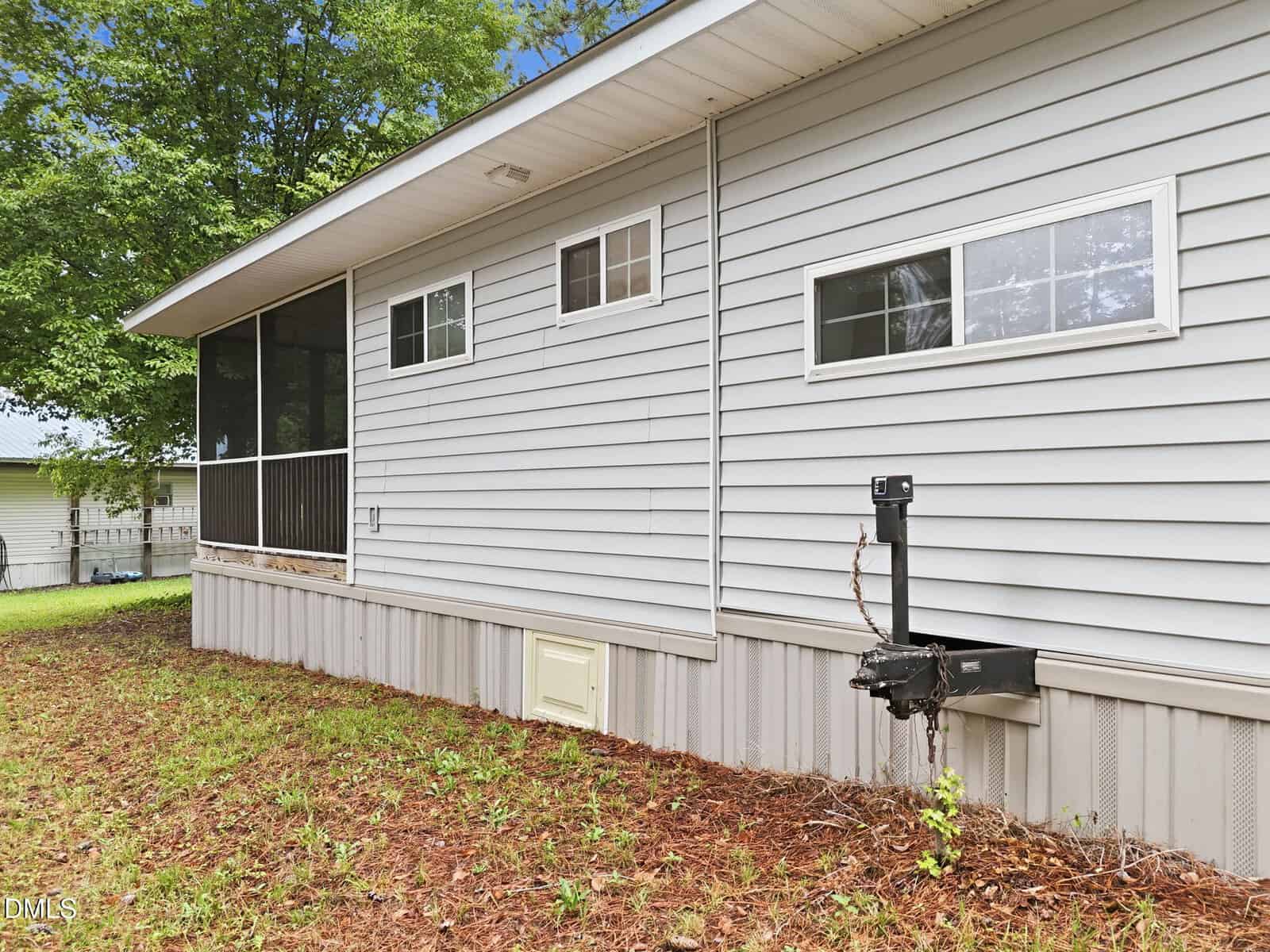 Side view of a light gray manufactured home with two small windows, screened porch, metal skirting, hitch at the front, grassy yard, and trees in the background on a cloudy day.