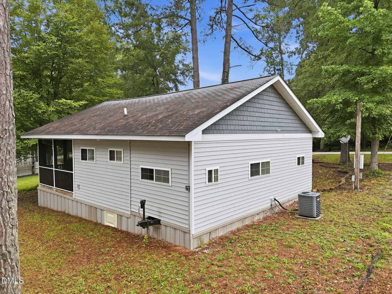 A small, single-story house with light gray siding and a dark shingled roof sits among tall trees on a grassy lot. It features several windows and a screened porch on one side, with an outdoor air conditioning unit nearby.