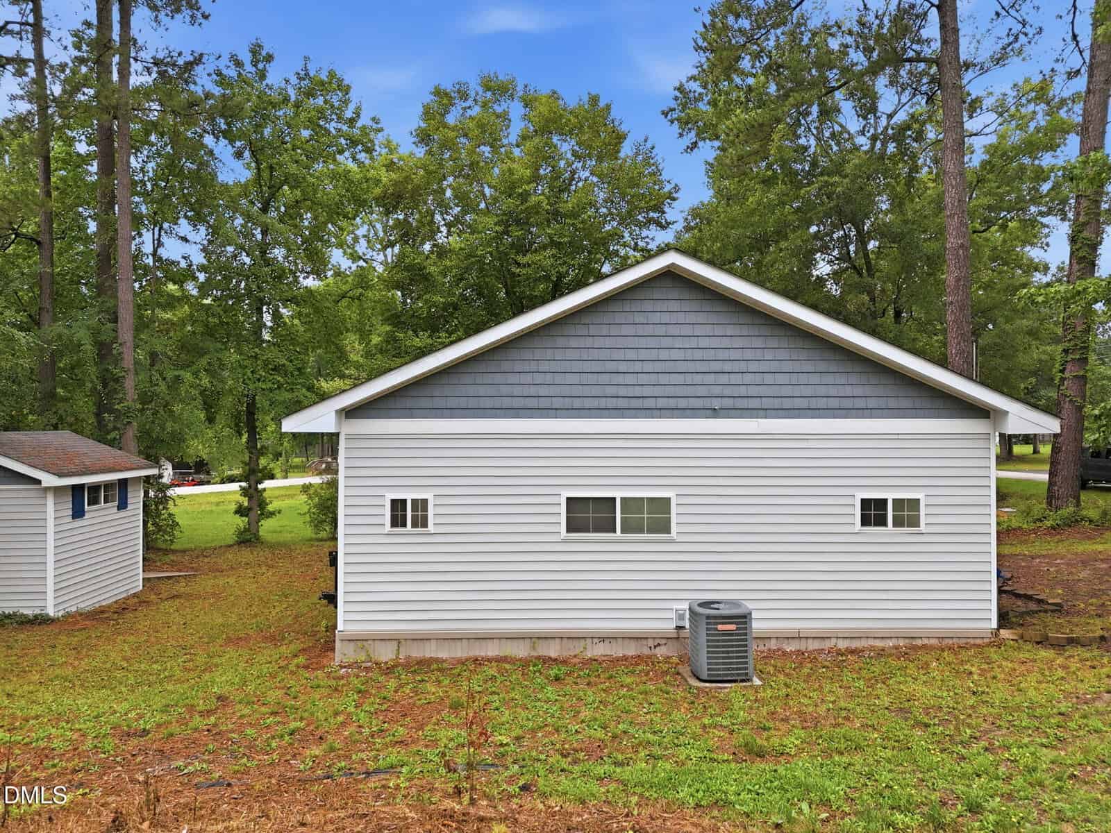 A gray and white house with a gable roof, horizontal siding, and small windows sits near a lawn and trees. An outdoor HVAC unit is next to the house, with a smaller shed visible on the left.