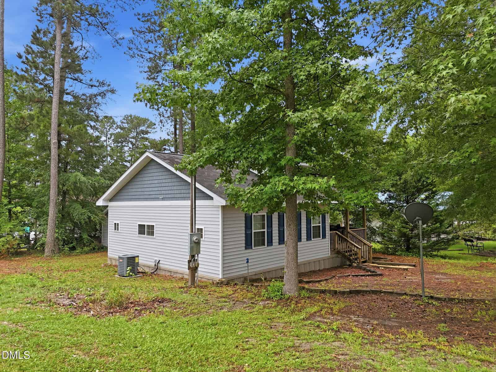 A small gray and blue house with white trim is surrounded by tall trees and green grass. A wooden porch is visible, along with a satellite dish and HVAC unit on the side of the house.