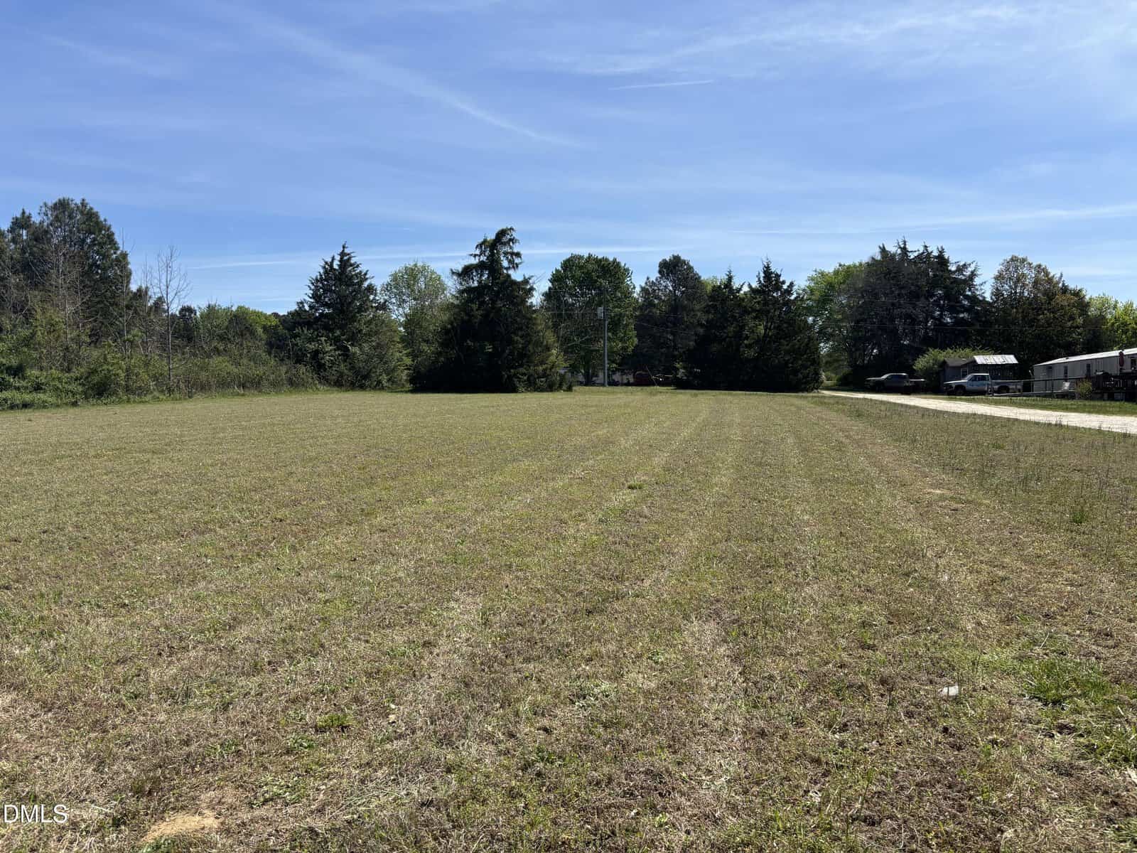 A large, open grassy field with trees and a clear blue sky in the background. There are some vehicles and a dirt road on the right side of the image.