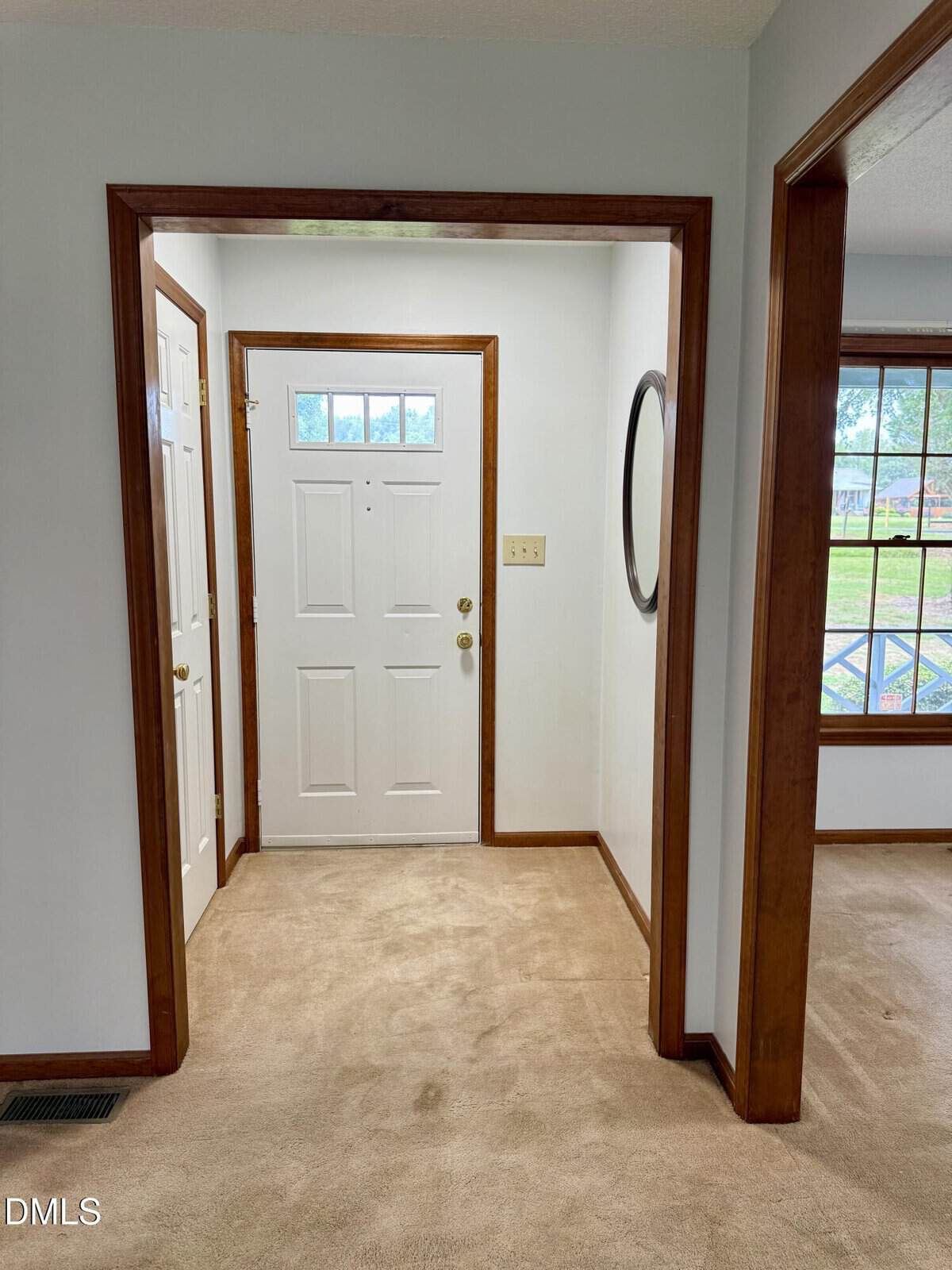 A hallway with beige carpet leads to a white front door with three small windows. The walls are light-colored with medium wood trim. There is a round mirror on the right wall and a window looking outside is visible to the right.