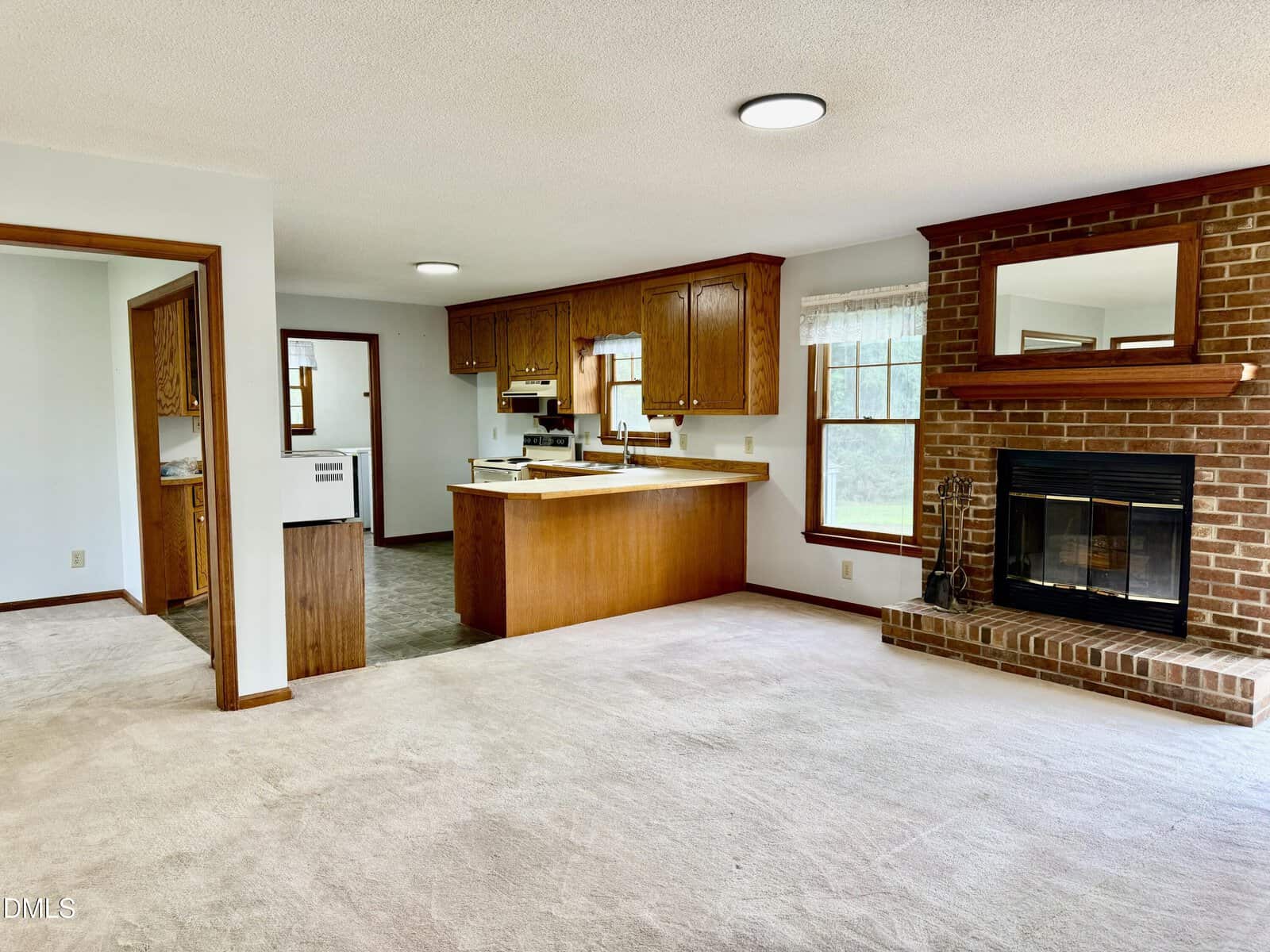 Open living area with light carpet, brick fireplace with mirror above, and a kitchen with wooden cabinets and white appliances. Large windows let in natural light.
