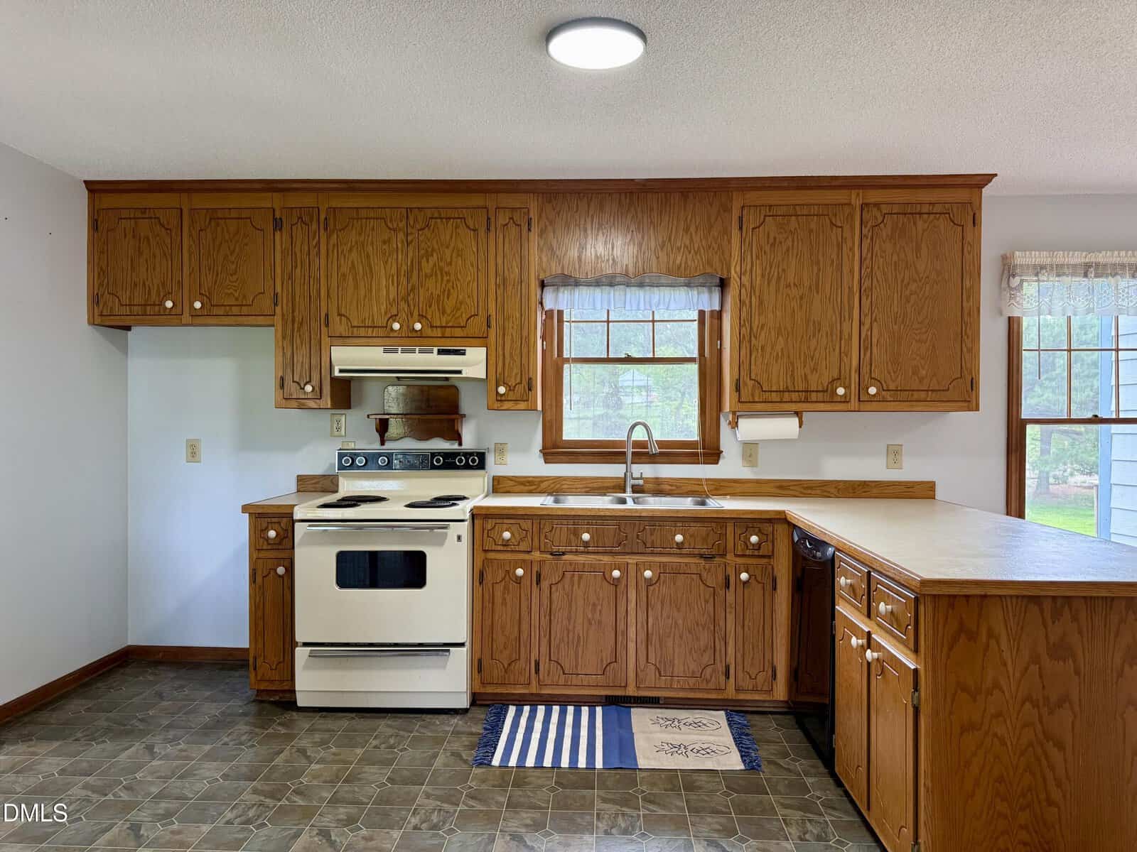 Kitchen with wooden cabinets, white stove, sink under a window with blue curtains, dishwasher, and tan countertops. Floor is dark patterned tile, and two small rugs are in front of the sink and stove.