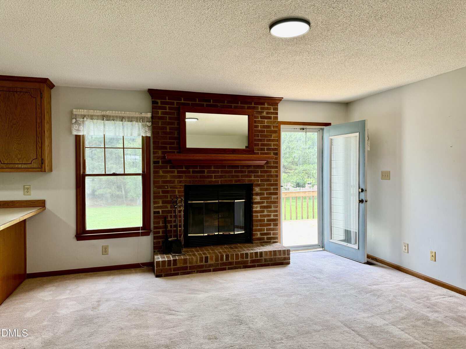 A living room with a brick fireplace, a large mirror above it, a window with a valance, a glass door leading outside, and beige carpet flooring. The room is bright with natural light.