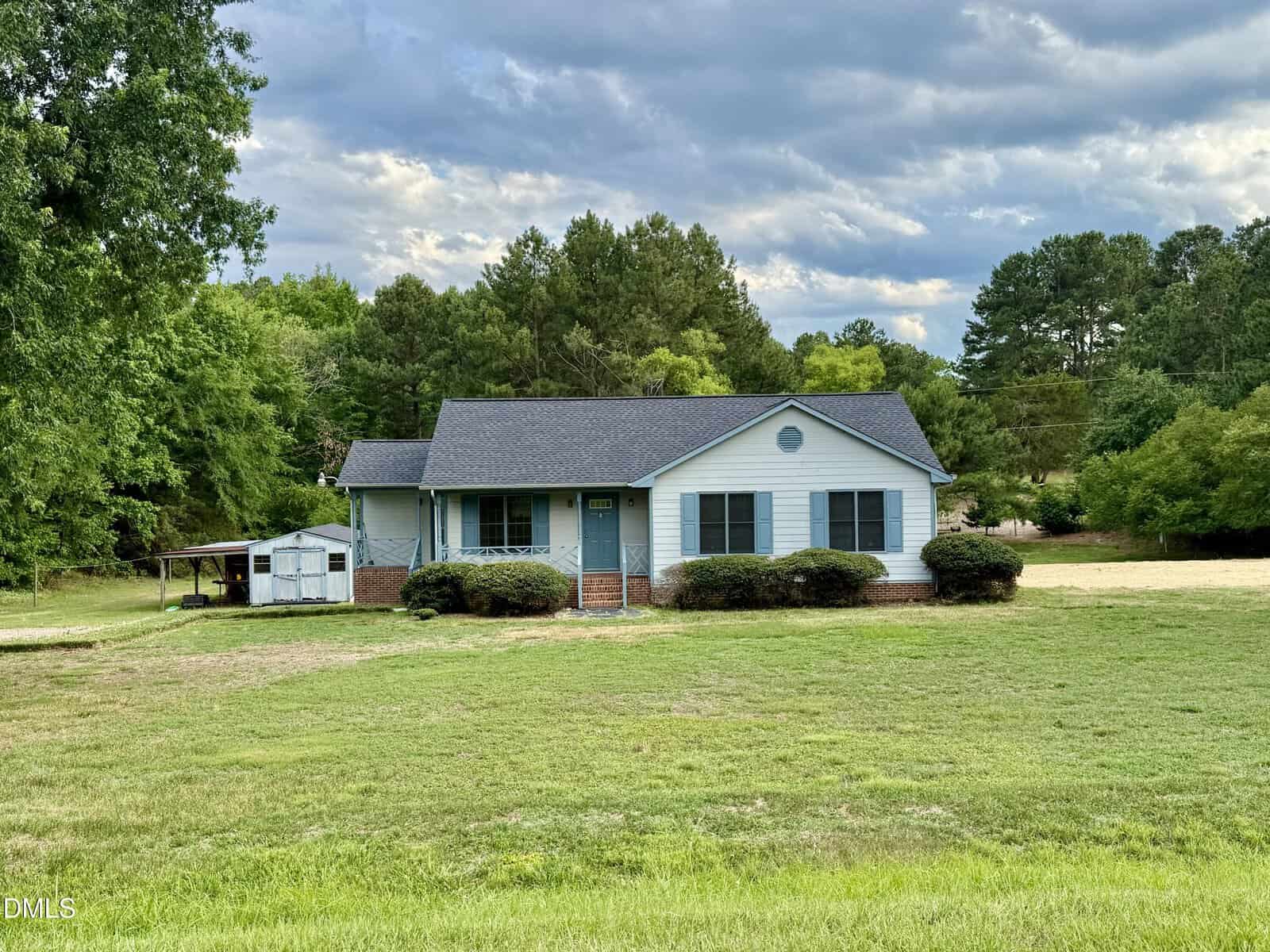 A light blue, single-story house with dark shutters sits on a large, grassy lawn at 460 Doctor Parrott Road in Henderson, surrounded by trees under a partly cloudy sky. A small shed is visible to the left of the house.