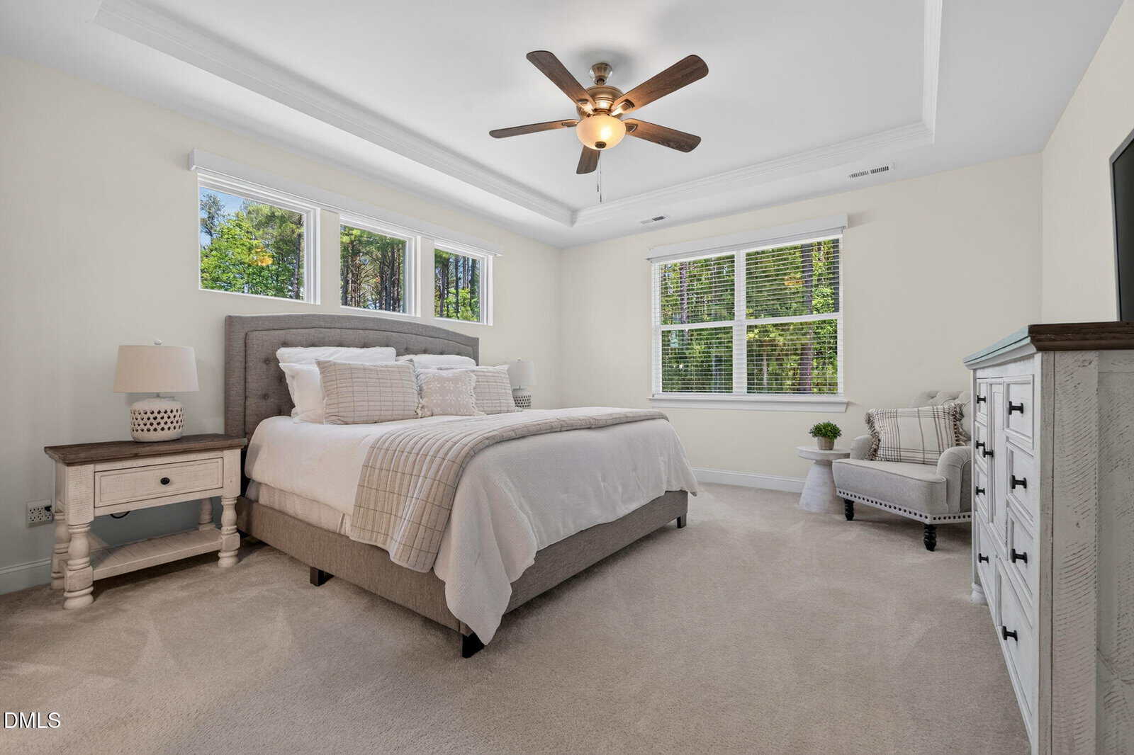 Bright bedroom at 75 Mallory Oak Drive in Franklinton features a large bed, neutral bedding, two nightstands, dresser, armchair, and big windows for natural light. Ceiling fan with light fixture and plush beige carpet complete the space.