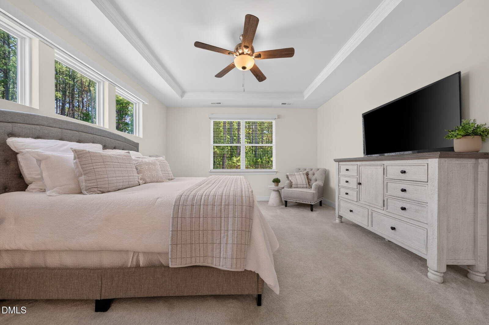 A spacious, bright bedroom with a large bed, neutral bedding, a ceiling fan, a dresser with a TV, an armchair by two large windows, and beige carpet flooring. Sunlight streams in, illuminating the room.