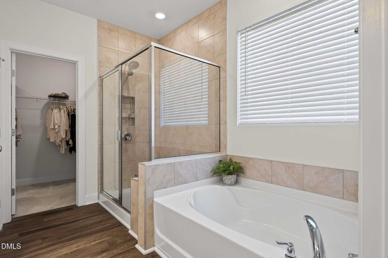 Modern bathroom with a glass-enclosed shower, a white bathtub with chrome fixtures, tan tile walls, a window with blinds, wooden flooring, and a small plant on the tub ledge; walk-in closet visible through open door.