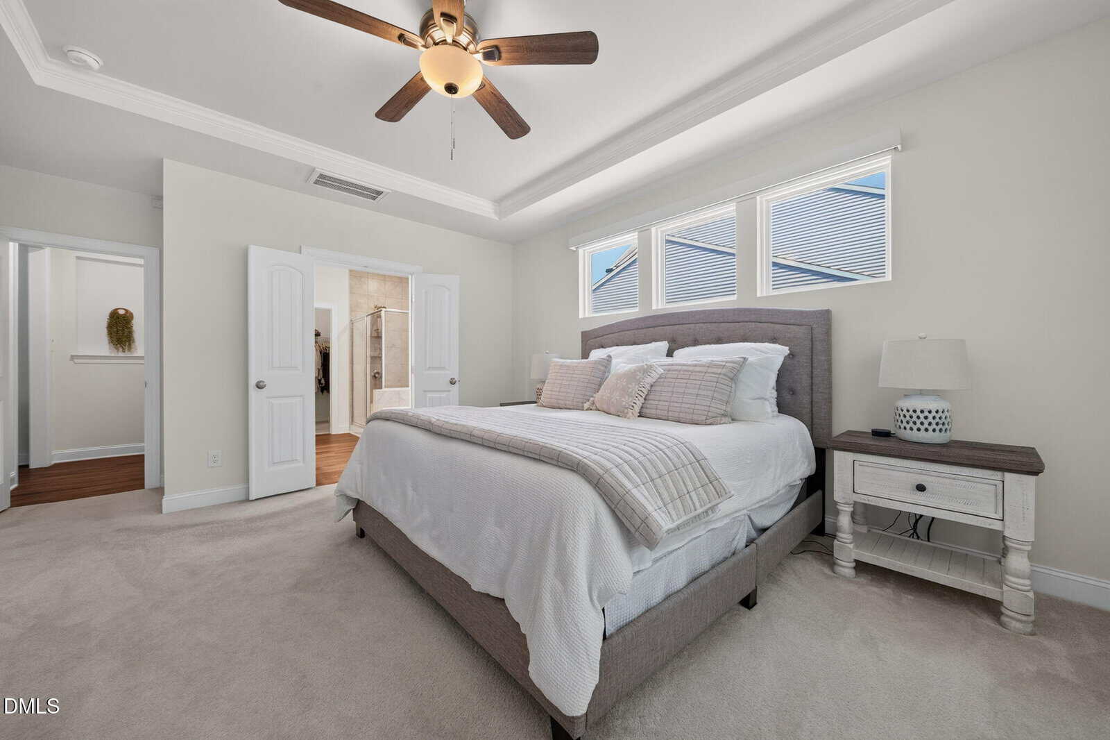 Bright bedroom with a gray upholstered bed, white bedding, two pillows, a nightstand with a lamp, ceiling fan, carpeted floor, and three windows. A door leads to an en-suite bathroom.