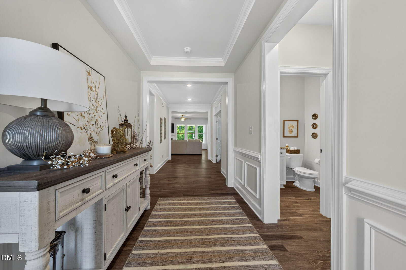 A bright hallway with wood floors, a striped rug, and white wainscoting. A console table with decor is on the left; an open doorway on the right leads to a bathroom. The hallway opens to a living room in the distance.