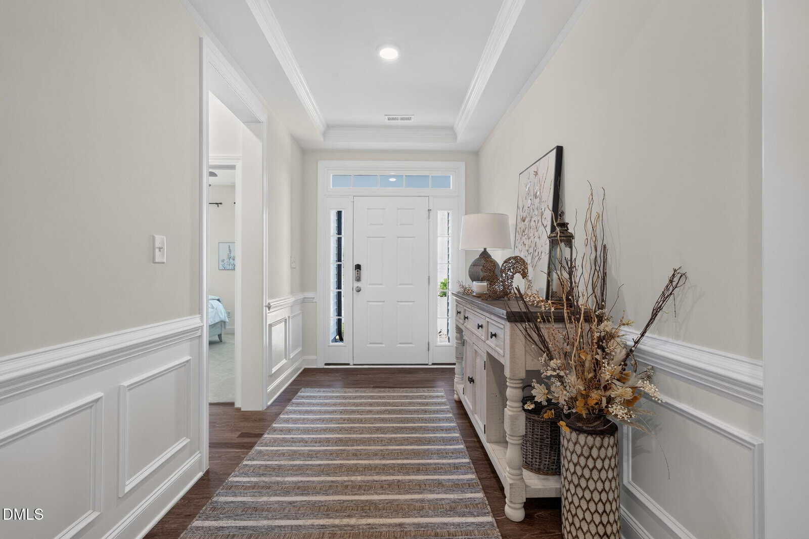 A bright entryway with a white front door, transom window, a console table with decorative items, a large woven basket with dried plants, and a striped rug on wooden flooring. White wainscoting lines the beige walls.