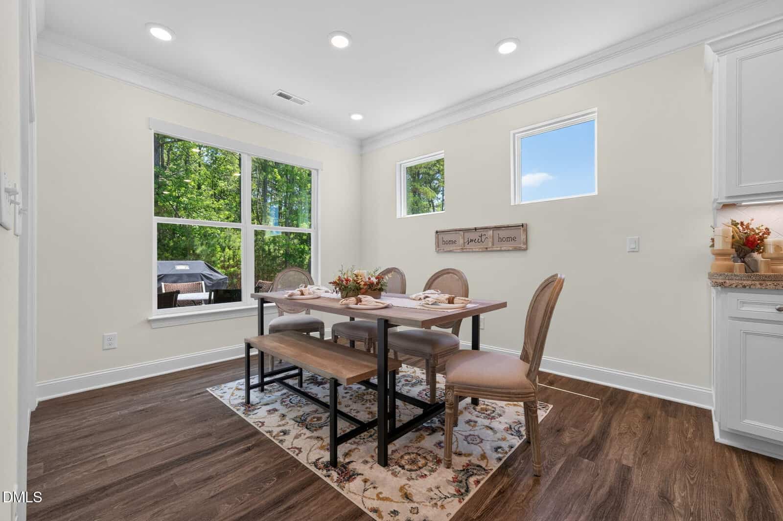 Bright dining area with large windows, a wooden table set for four, beige upholstered chairs, and a patterned rug on dark wood flooring. Wall decor with “home, family” hangs above the table. Trees visible outside.