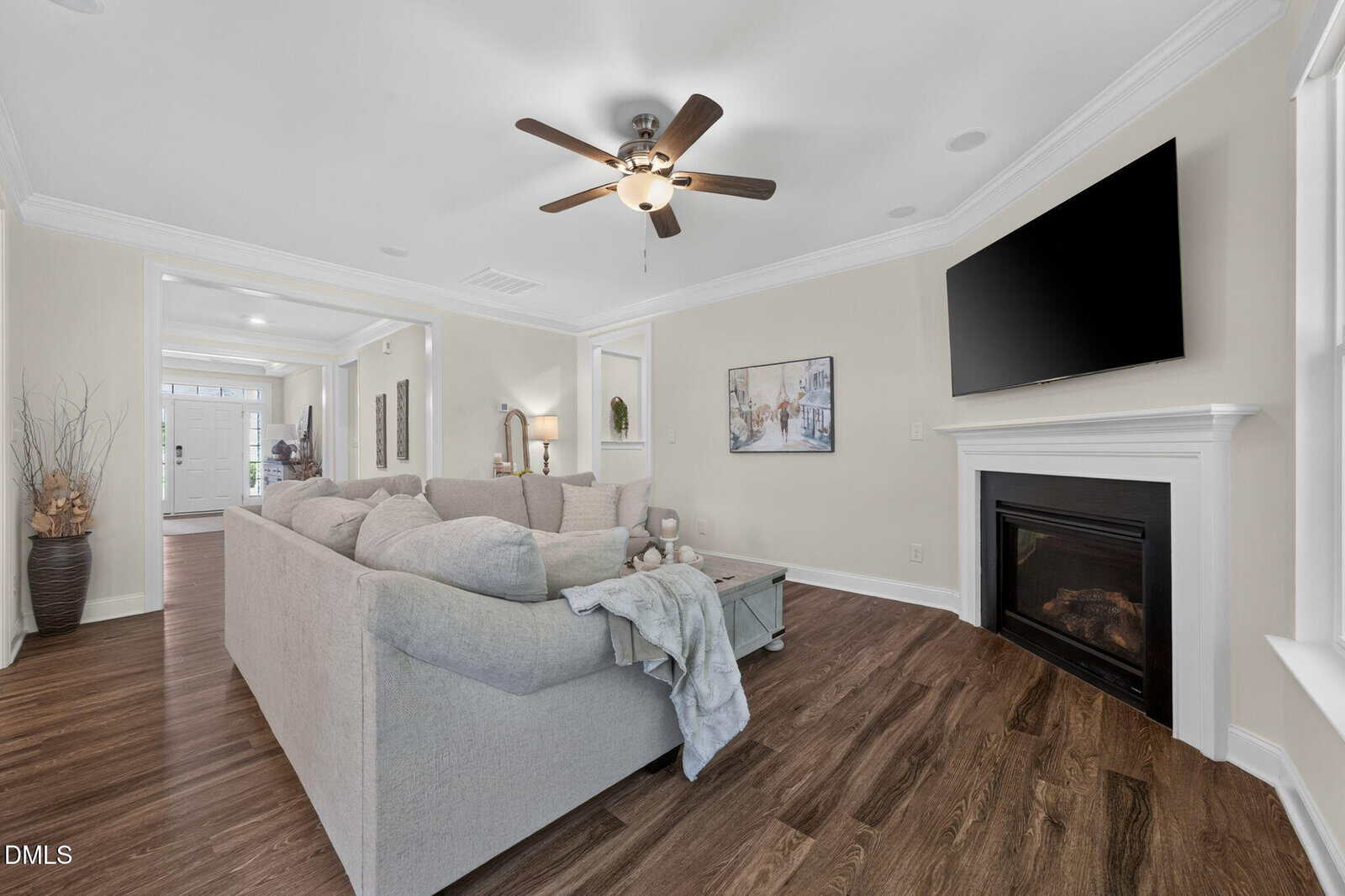 Bright living room with a gray sectional sofa, wooden floors, a ceiling fan, wall-mounted TV above a fireplace, and decor accents. Entryway and additional rooms visible in the background.