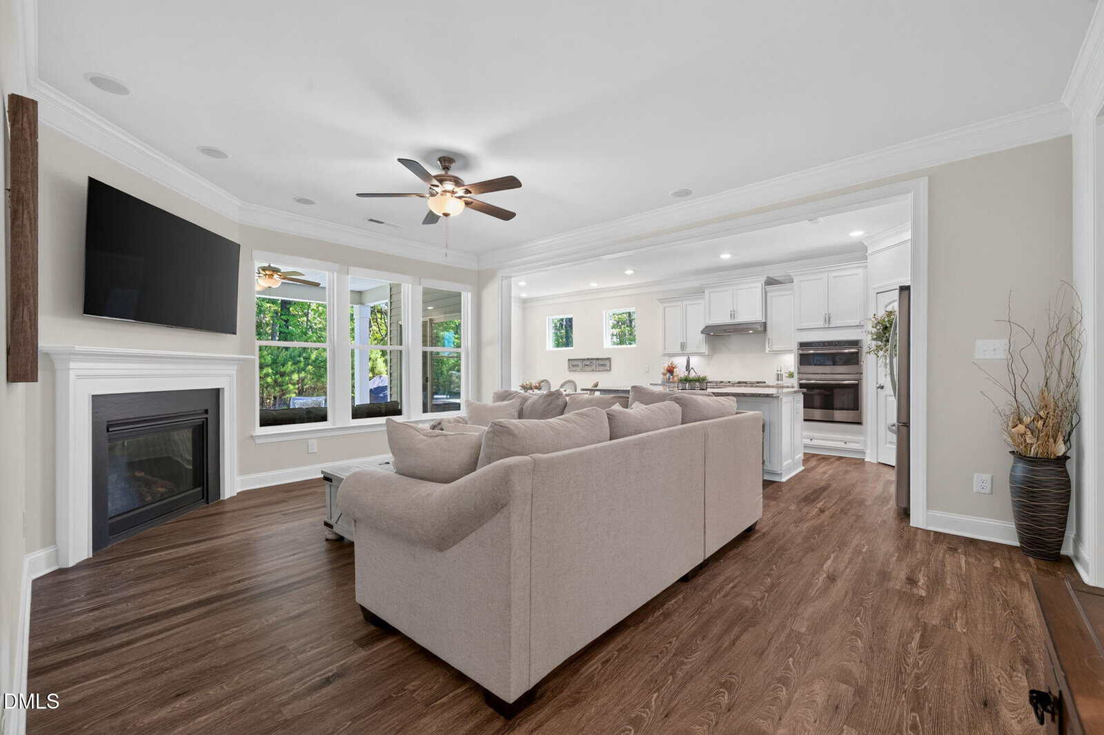 Spacious living room with beige sofas, wood floor, wall-mounted TV above a fireplace, large windows, and open view into a white kitchen with stainless appliances. Ceiling fan and recessed lights present.
