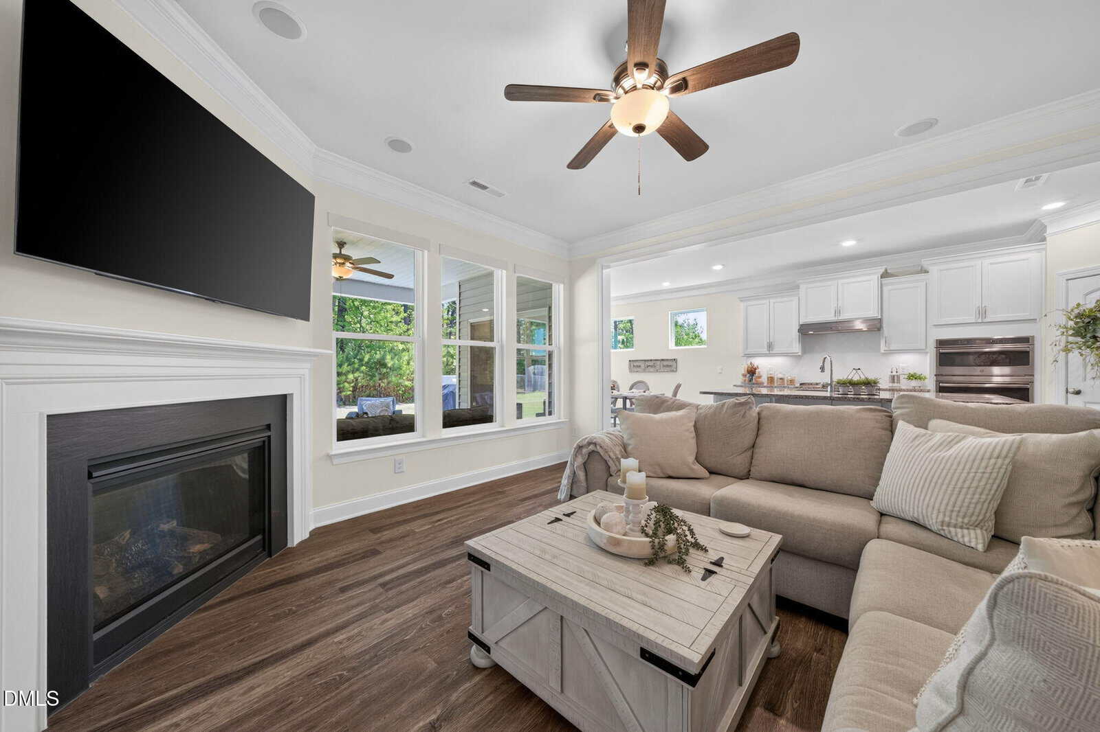 A bright, modern living room with a ceiling fan, mounted TV above a fireplace, beige sectional sofa, wooden coffee table, and large windows overlooking greenery, connected to a white kitchen in the background.