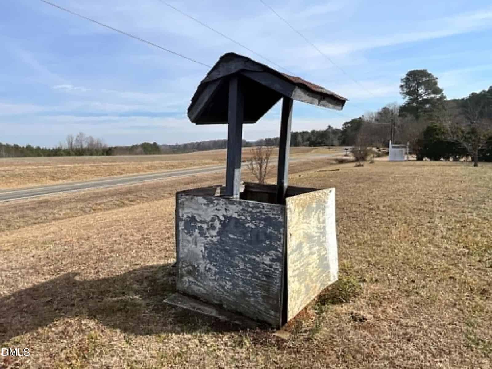 A weathered, wooden well with a slanted roof stands in an open grassy field near a road, with trees and a clear sky in the background.