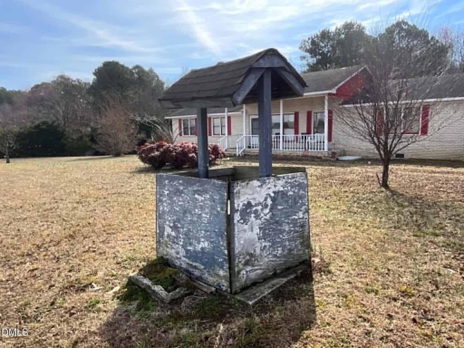 A weathered wooden wishing well stands in a grassy yard with a single-story house, bushes, and trees in the background under a partly cloudy sky.