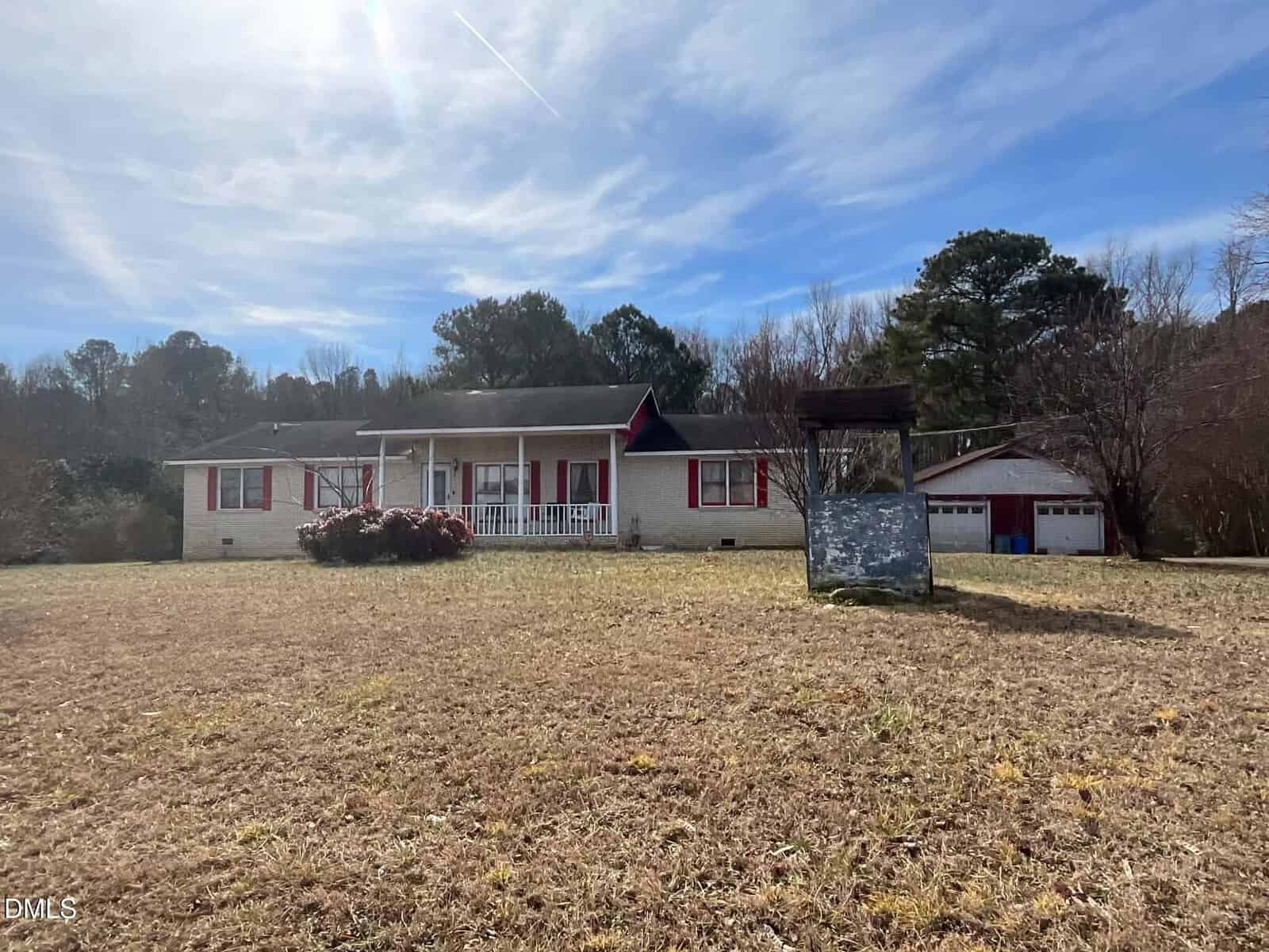 A ranch-style house with red trim and shutters sits in a dry, grassy yard. A detached garage and an old well are also on the property under a bright, cloudy sky.