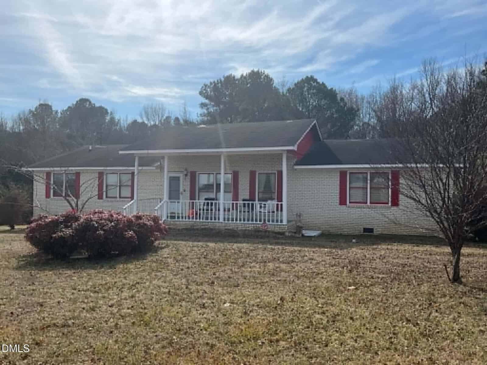 Charming single-story home at 44 Sutton Road, Louisburg, with light-colored brick, red shutters, and a cozy covered front porch. A bare tree and bush grace the front yard, framed by trees under a partly cloudy sky.