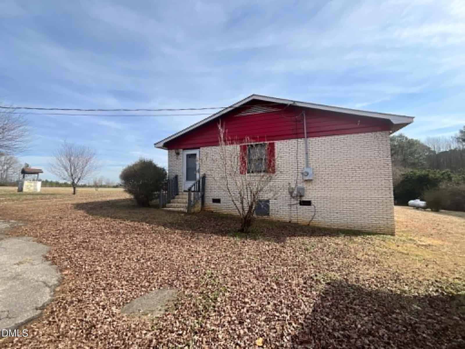 A small single-story house with light brick walls and a red gable roof sits on a lot covered with dry leaves. There are a few shrubs near the entrance and leafless trees in the background under a partly cloudy sky.