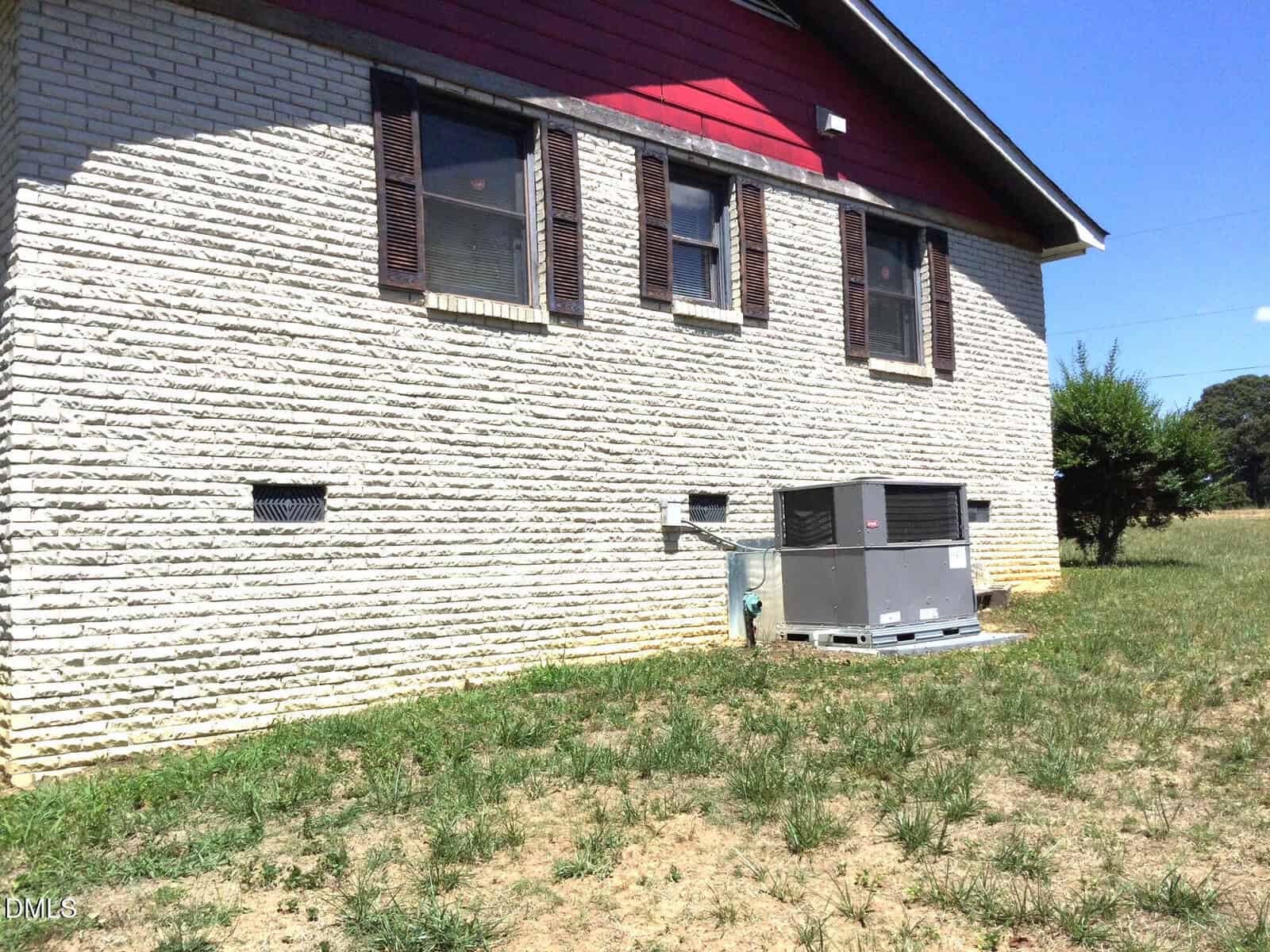 A brick house with three windows and brown shutters has an air conditioning unit installed outside on a concrete pad. The yard has patchy grass and the sky is clear.
