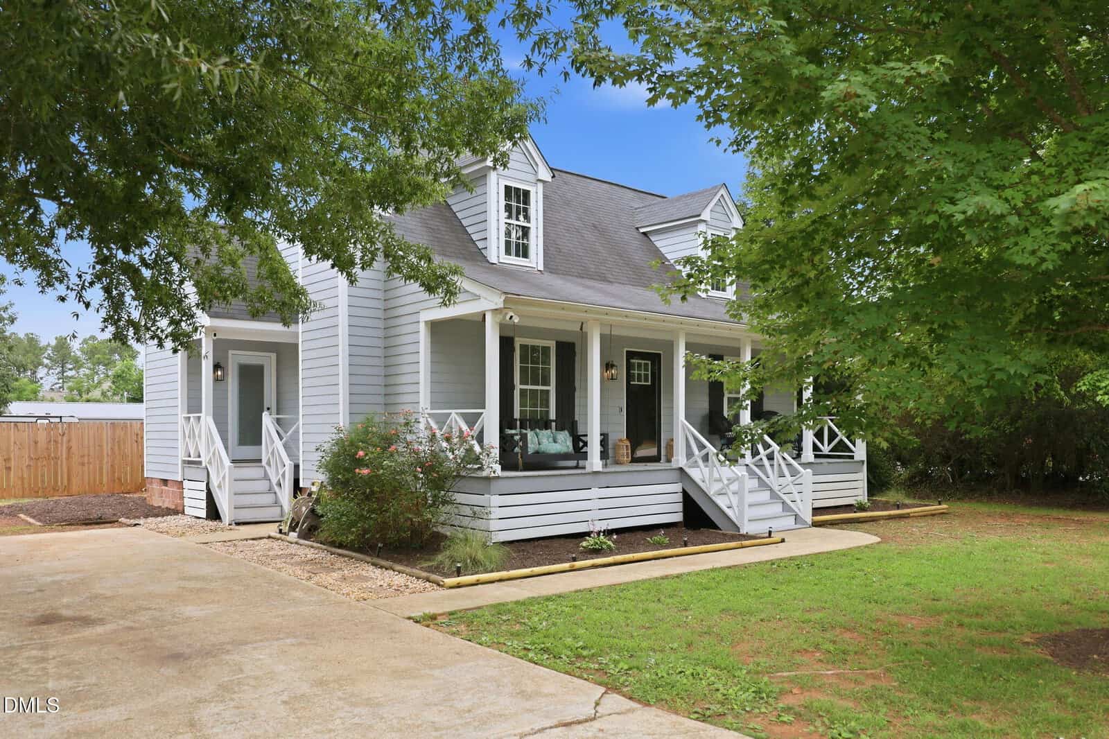 A light gray two-story house with white trim, black shutters, and a covered front porch with seating. Large trees and a driveway frame the neatly landscaped yard.