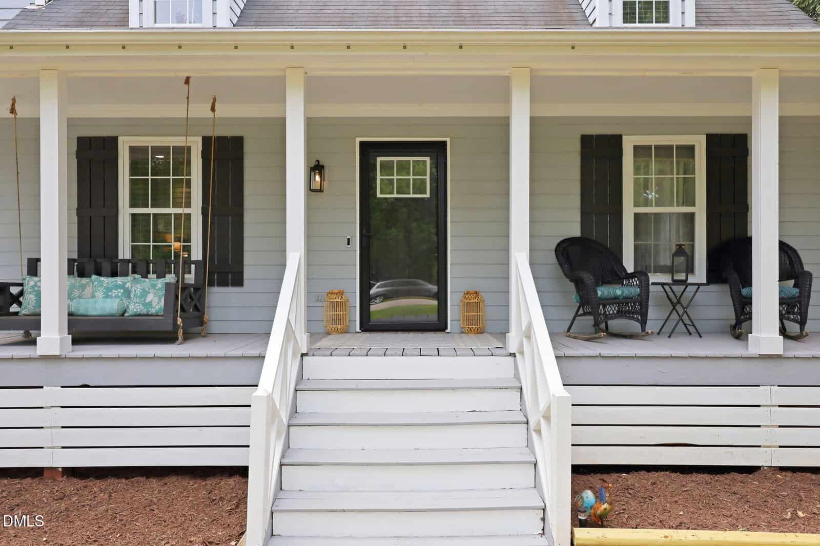 White house front porch with gray steps, white railings, two black rocking chairs on the right, a black porch swing with teal cushions on the left, and two lanterns by the black front door.