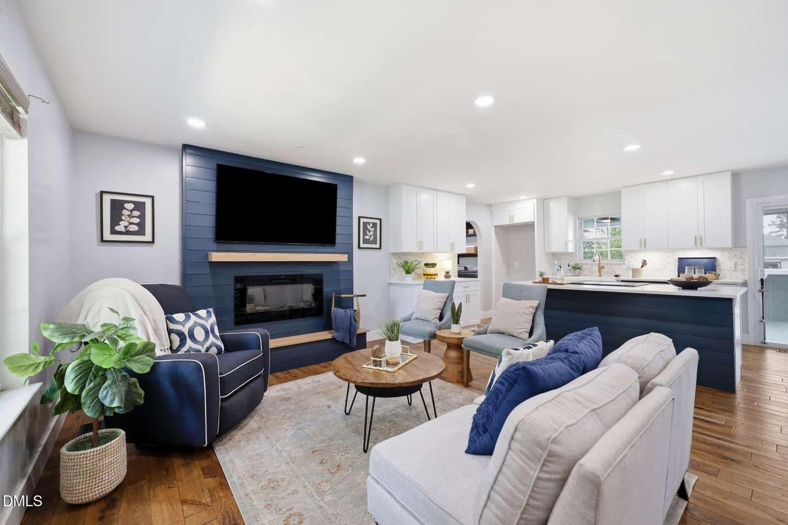 A modern living room with white walls, navy blue accents, a mounted TV above a fireplace, light sofas with blue pillows, a round coffee table, hardwood floors, houseplants, and an open view to a bright kitchen.