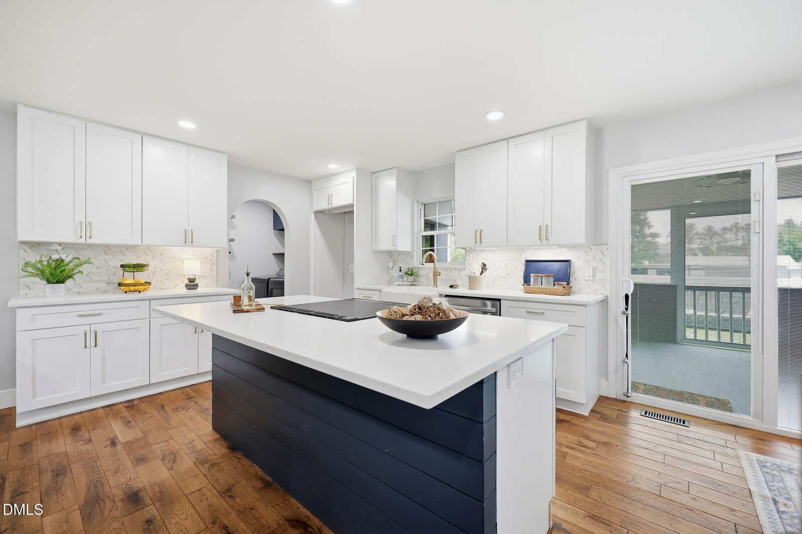 Modern kitchen with white cabinets, white countertops, a navy island, wood floors, and a sliding glass door leading to a screened porch. Decorative items and plants are on the counters. Bright and spacious atmosphere.