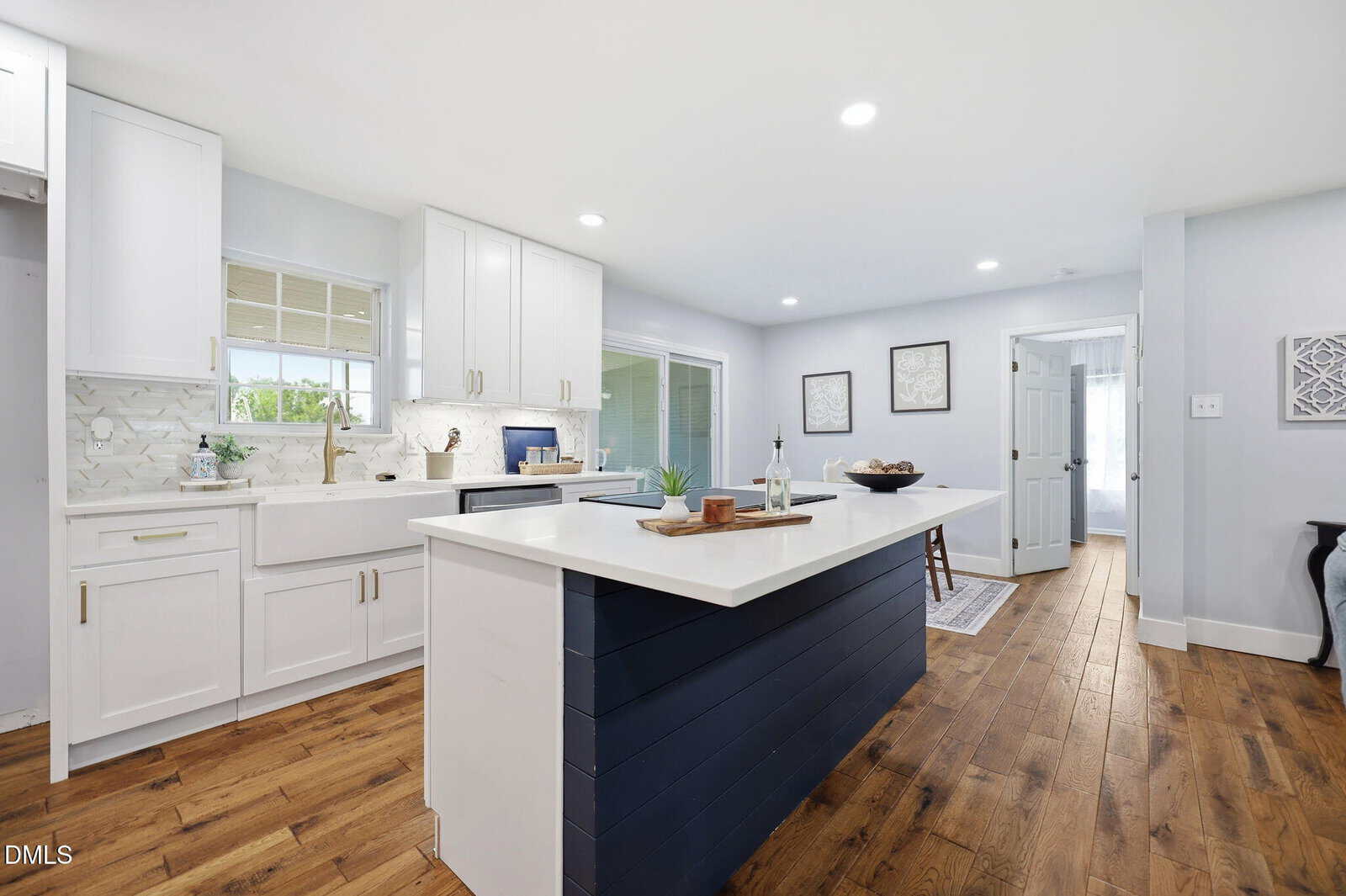 Modern kitchen with white cabinets, farmhouse sink, white countertops, navy blue island, wood flooring, and geometric backsplash. Bright space with recessed lighting and decor accents, leading to a hallway and dining area.