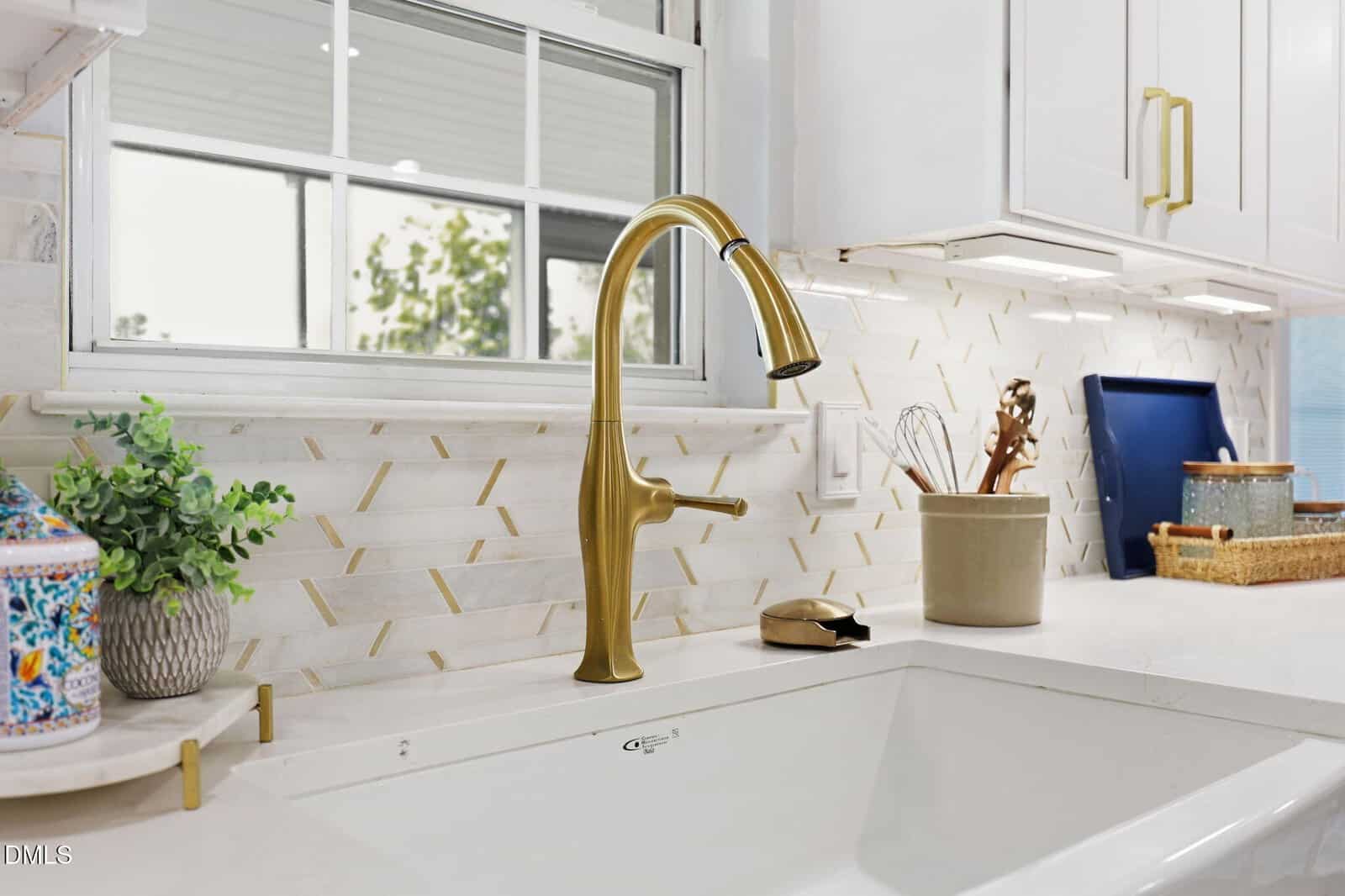 A modern kitchen sink with a gold faucet, white countertops, beige backsplash with gold accents, potted plant, utensils in a beige holder, and a blue tray near a window with a view of greenery outside.