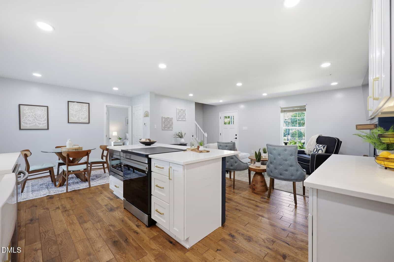 Bright open-concept kitchen and living area with wood floors, white cabinets, a center island, dining table, and light blue armchairs near windows; modern and inviting decor.