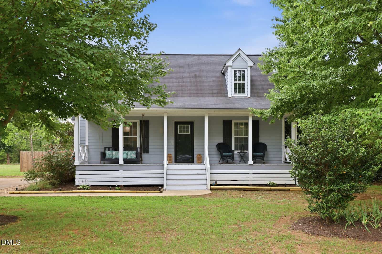 A light gray house at 100 New Castle Court in Youngsville with a covered front porch, black shutters, white trim, and steps leading to the front door. The porch features chairs and is surrounded by green trees and lawn.