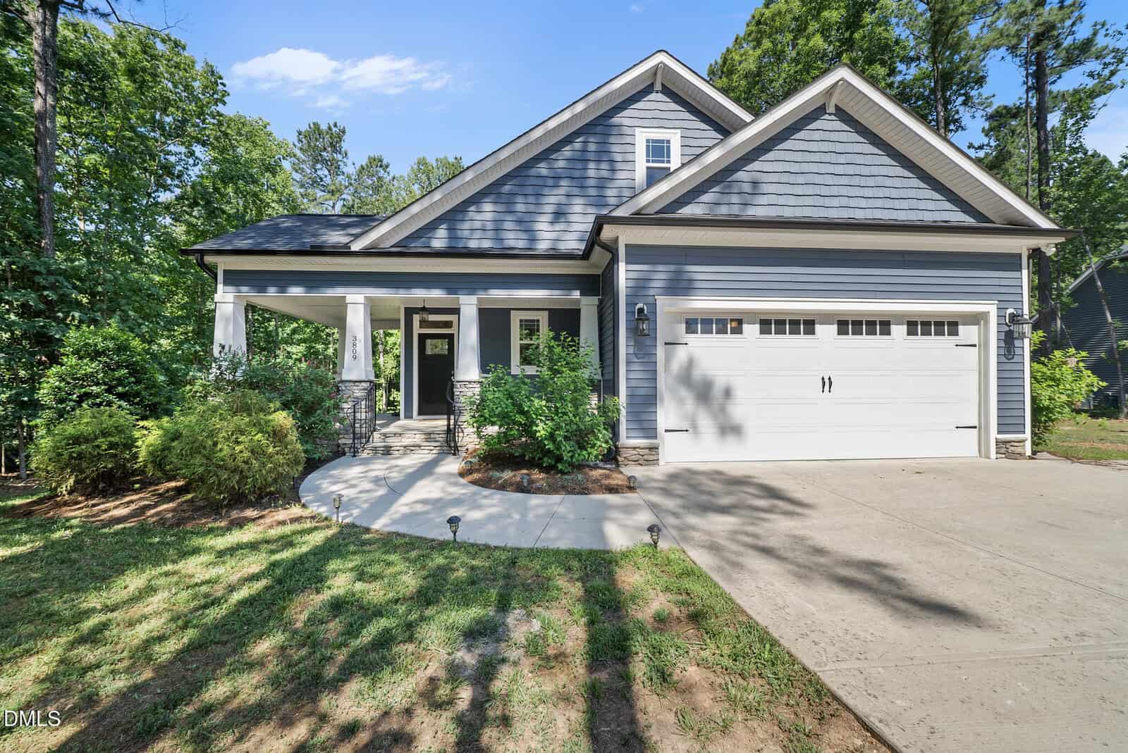 A modern, single-story house at 3809 Watermark Drive, Franklinton, features gray siding, a covered front porch, white trim, and a double garage. A concrete driveway curves to the porch amid green shrubs and tall trees in the background.