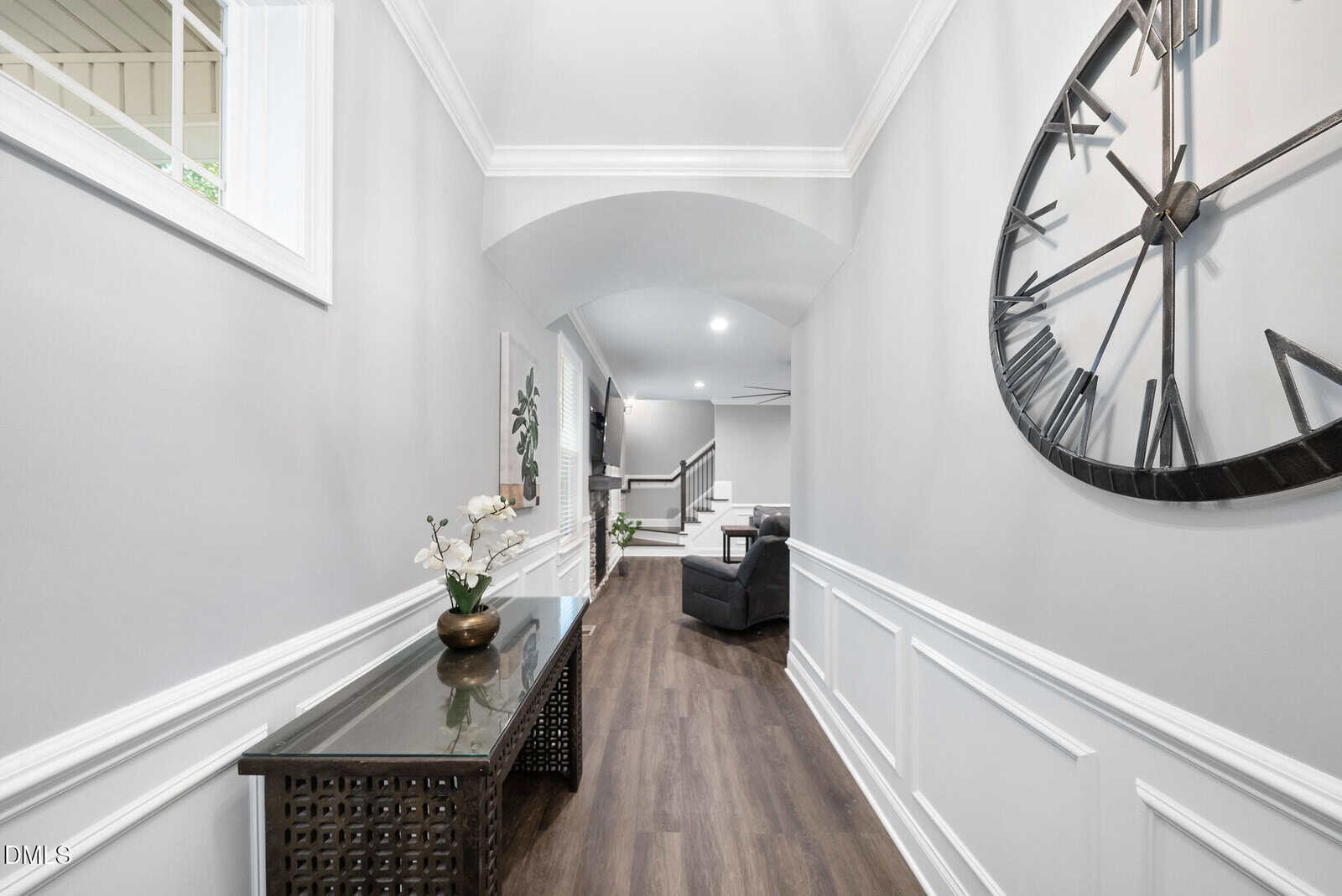 A bright hallway with white wainscoting, light gray walls, a dark wood console table with a potted orchid, and a large decorative wall clock, leading to a living area with seating.