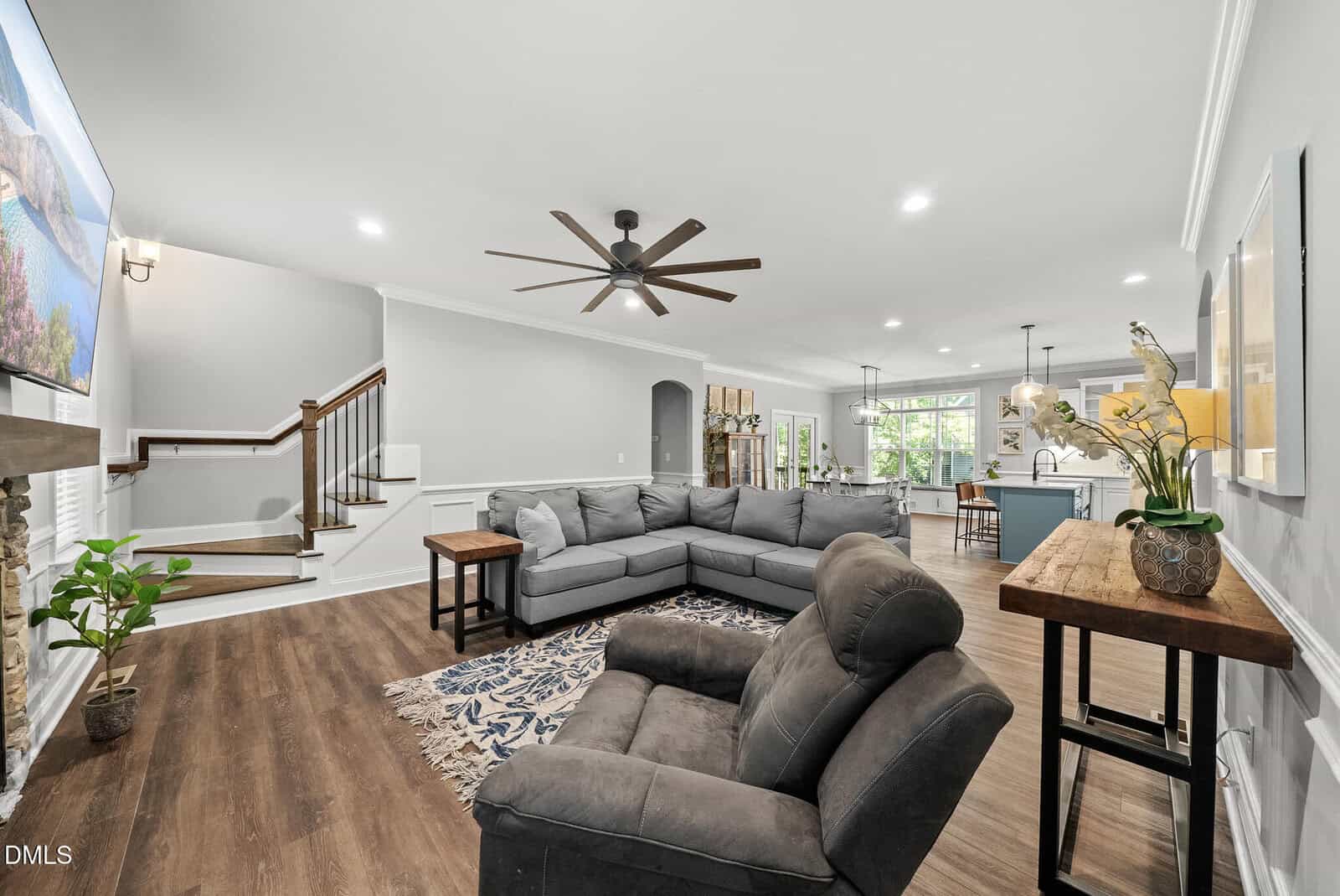 Modern living room with gray sectional sofa, armchair, wooden floors, and a large ceiling fan. The space is open to a bright kitchen and dining area, with stairs leading upstairs on the left. Plants and decor add warmth.