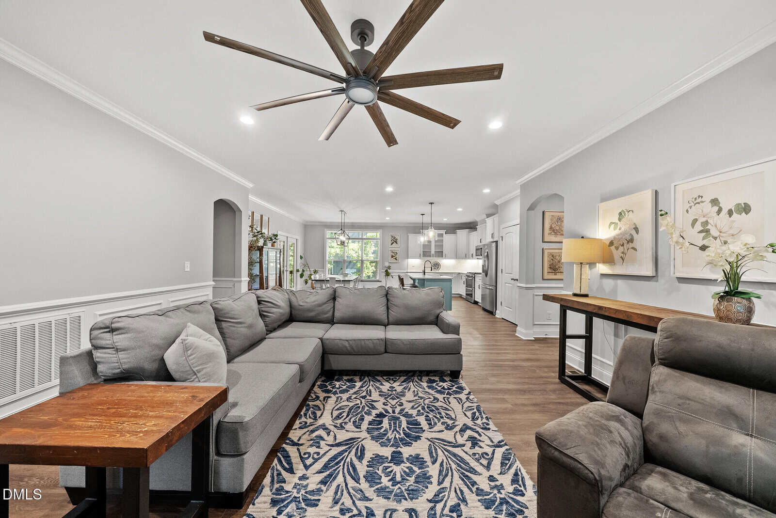 A spacious living room with a large gray sectional sofa, patterned blue and white rug, wooden side tables, and a ceiling fan. The open floor plan extends to a kitchen with white cabinets and modern lighting.