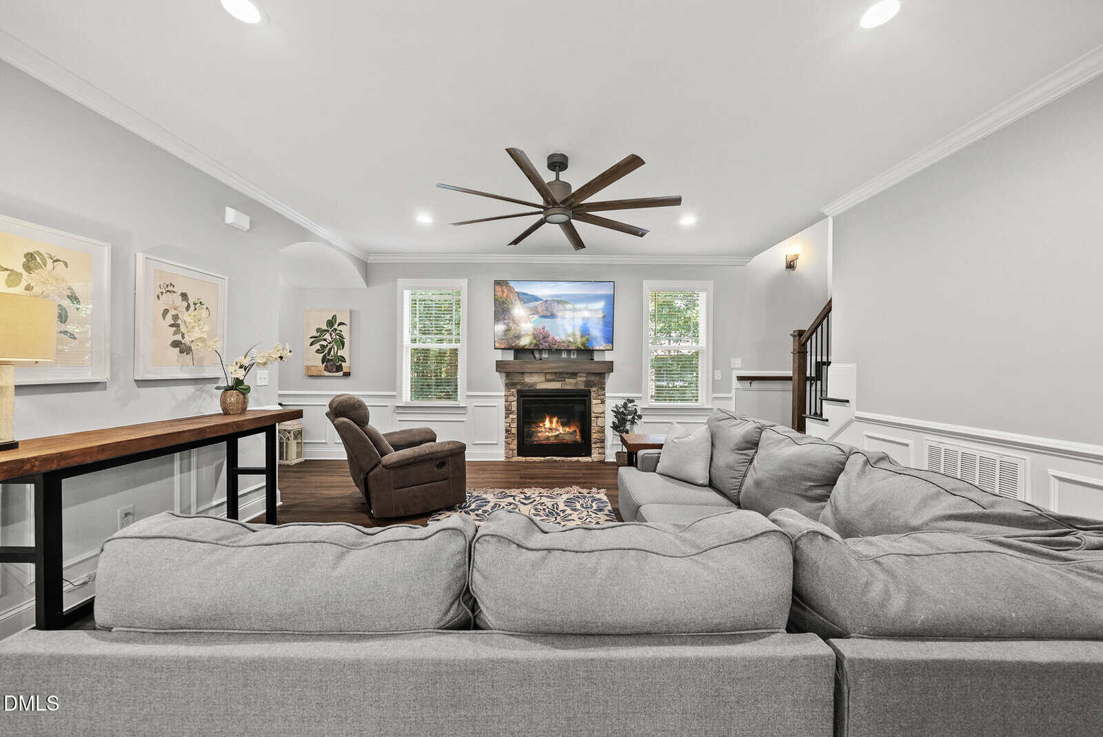 Modern living room with a large gray sectional sofa, fireplace with TV above, armchair, desk with lamp and orchid, artwork on the walls, ceiling fan, and staircase in the background.