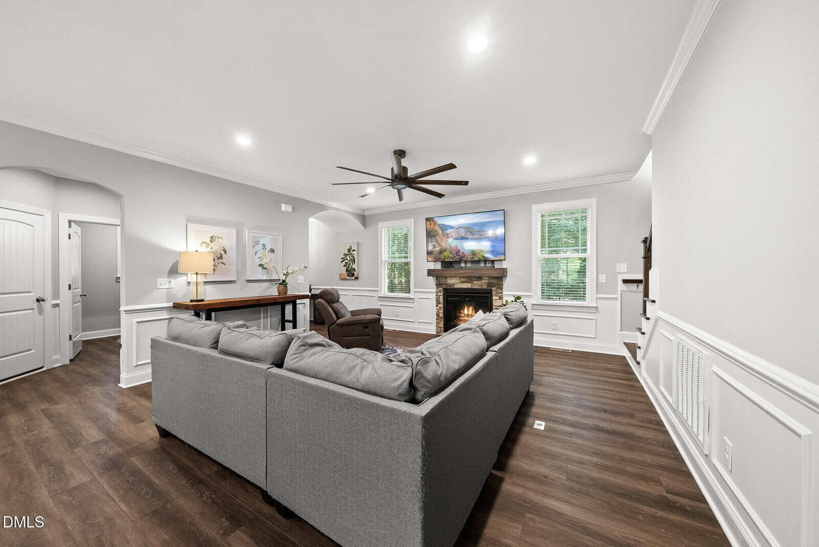 Bright living room with a gray sectional sofa, armchair, fireplace, wall-mounted TV, wood floors, white trim, large windows, ceiling fan, and a console table with a lamp and artwork.