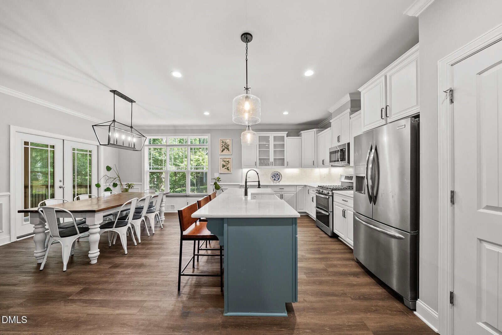 Modern kitchen with white cabinets, stainless steel appliances, a large island with teal base, two pendant lights, and a wooden dining table with white chairs by large windows. Warm wood floors throughout.