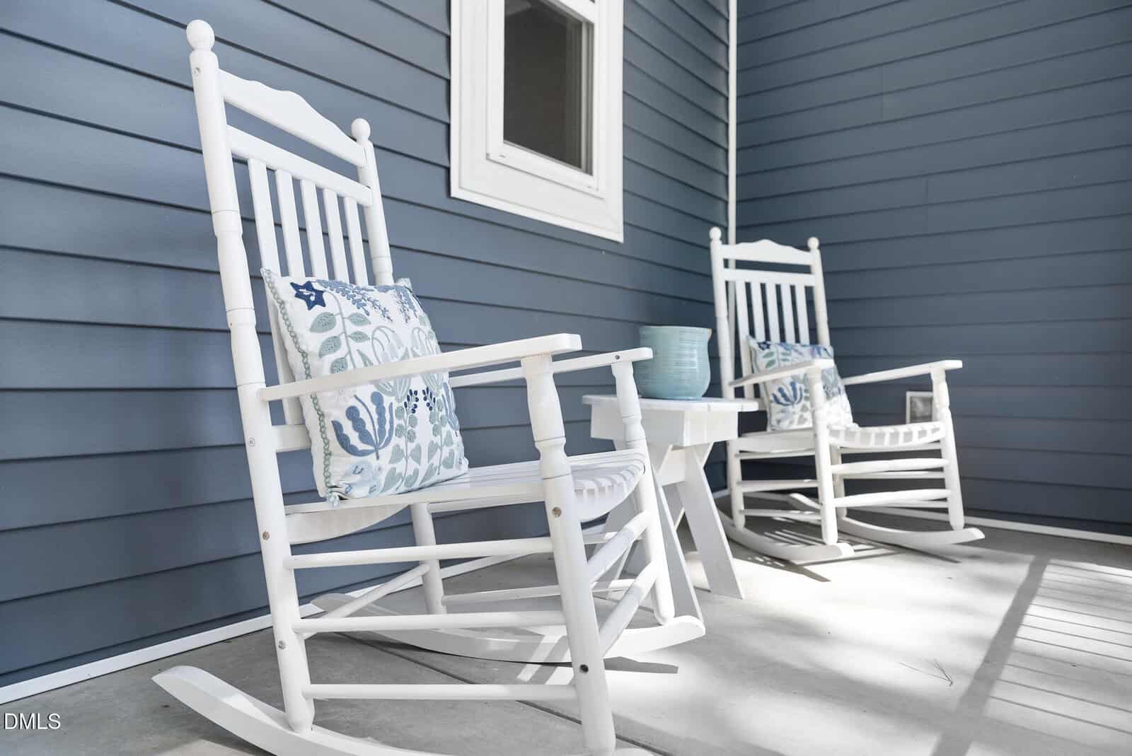 Two white wooden rocking chairs with patterned blue and white cushions sit on a porch with blue siding. A small white table between them holds a light blue vase and cup. Sunlight casts shadows on the concrete floor.
