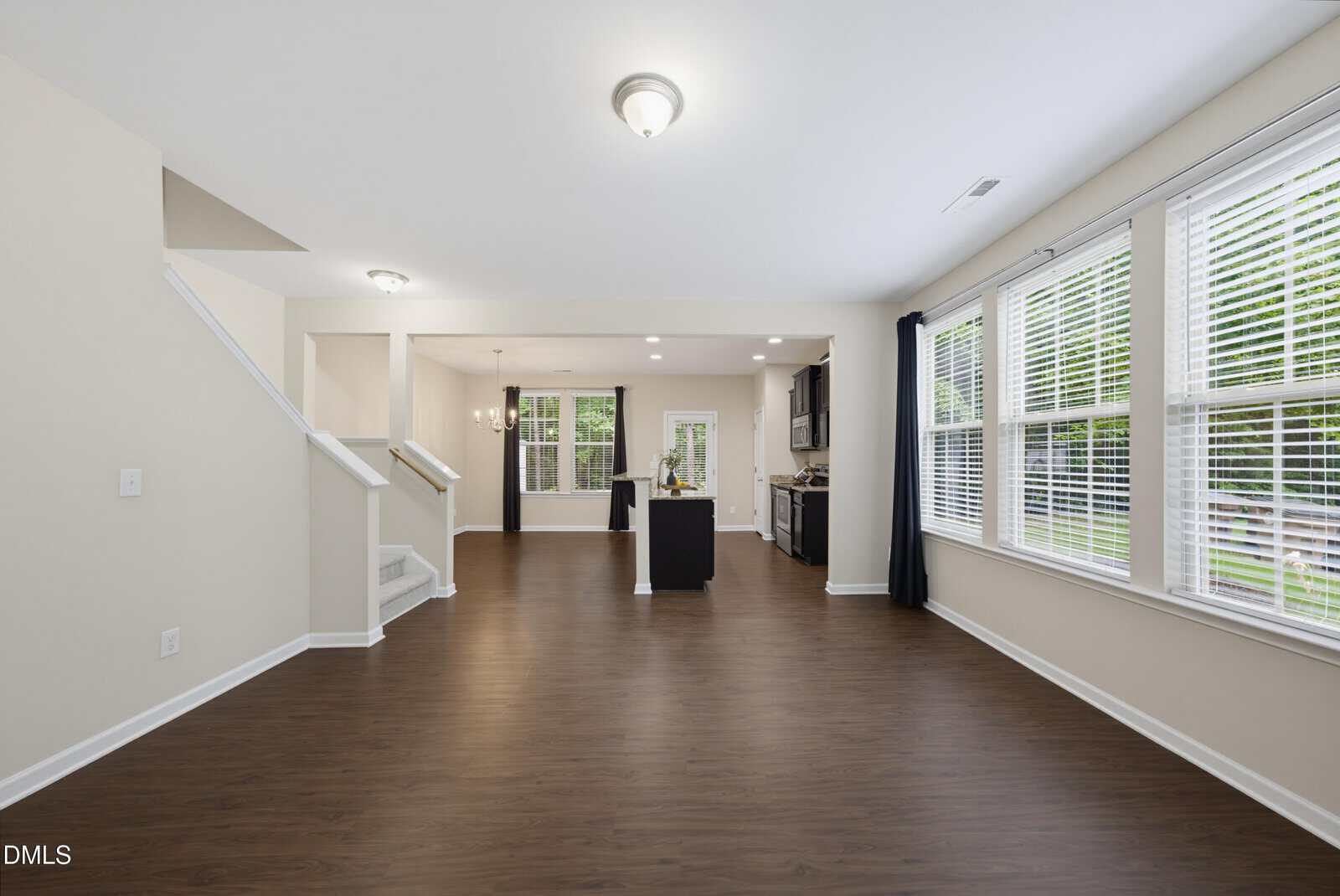 Open-concept living area with wood flooring, white walls, stairway on the left, dining area, modern kitchen, and large windows with blinds offering views of the backyard.