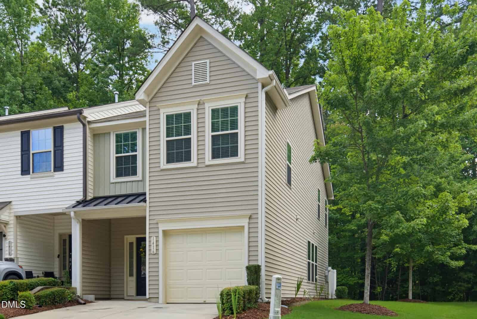 A beige two-story townhouse with white trim, a one-car garage, and multiple windows is surrounded by green trees and a well-kept lawn under a bright sky.