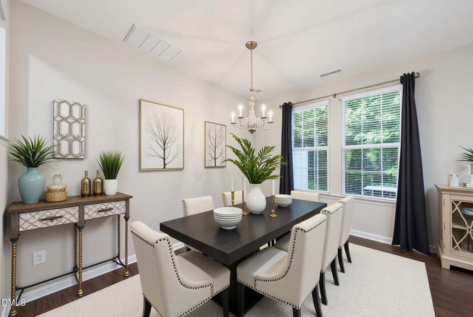 A modern dining room with a black table, eight cream-colored chairs, potted plants, wall art, a chandelier, and large windows with dark curtains letting in natural light.