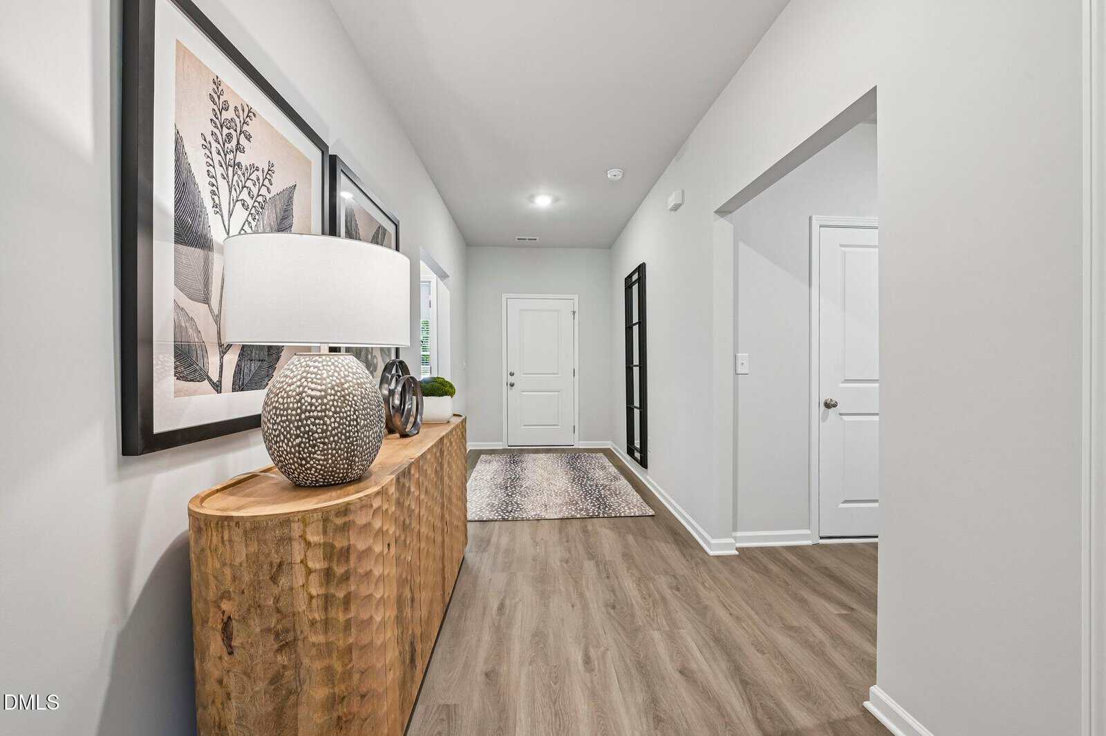 A hallway with light wood flooring, a textured wooden console table with a lamp and decor, framed art on the wall, a rug near the white front door, and white doors on the right side.