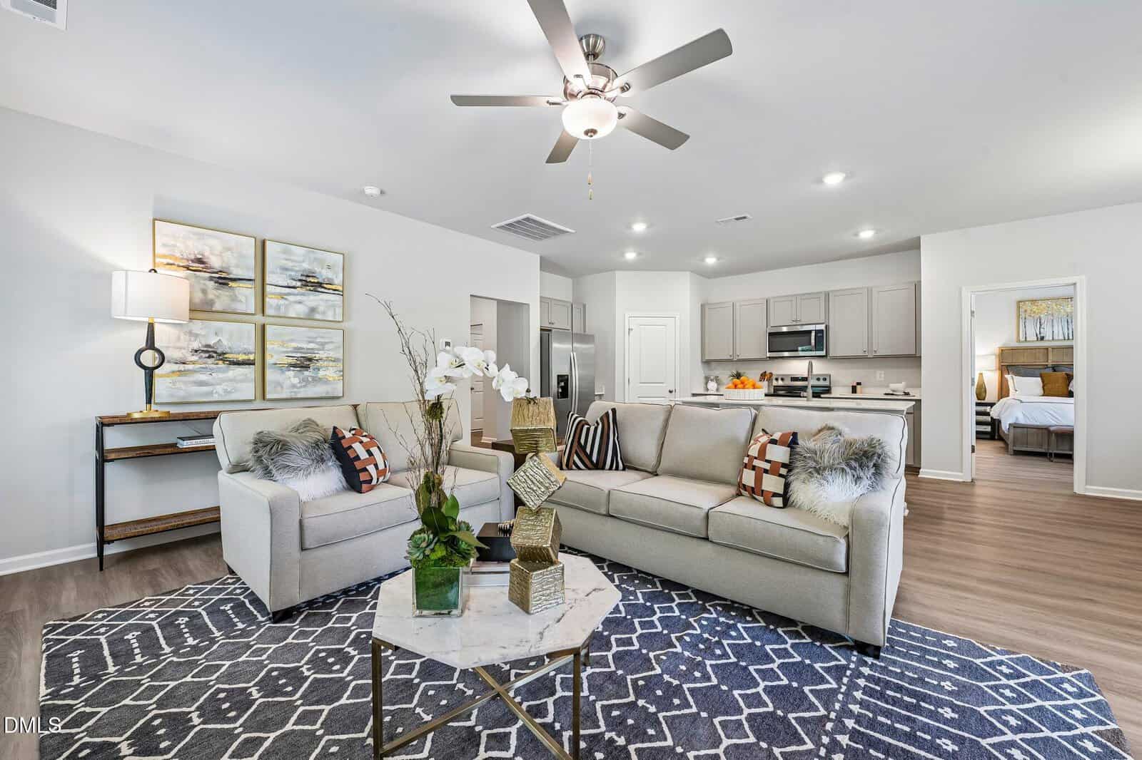 Modern living room with two beige sofas, a hexagonal marble coffee table, and a patterned rug. The space is open to a kitchen with light cabinets. Wall art, decorative pillows, and plants add accents. A bedroom is visible in the background.