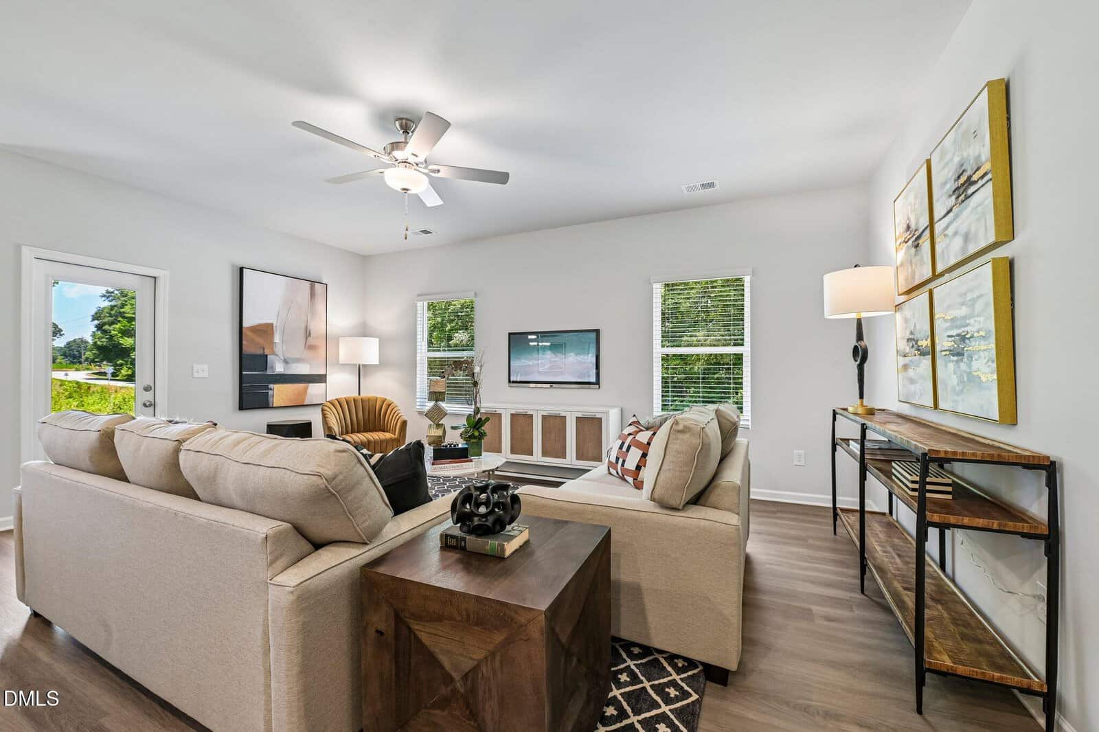 Living room with two beige sofas, a wooden side table, wall art, floor lamp, ceiling fan, and a TV mounted above a cabinet. Three windows and a glass door let in natural light. Decor includes books, plants, and framed paintings.