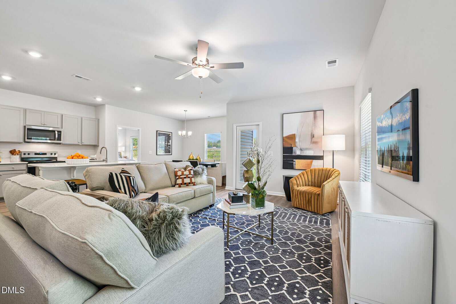 Modern living room with light sofas, patterned rug, and yellow armchair. Kitchen with stainless steel appliances is visible in the background. Walls are decorated with framed art, and a ceiling fan is centered above.
